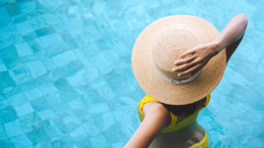 Tayland 'da yaz tatili konseptiyle seyahat ediyorlar. Asyalı kadının arka ve üst görüntüsü yüzme havuzu spa merkezinde rahatlıyor. Havuz başında hasır şapka ve bikiniyle oturuyor..