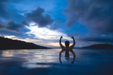 Tatil konseptiyle yaz seyahati. Asyalı adamın kollarını esnettiği arka manzara gün batımında açık havuz tatil köyü deniz manzarası. Yerli Doğa Turizmi Phuket Adası, Tayland