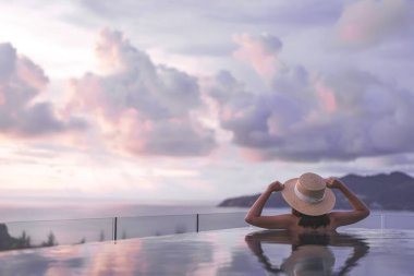 Güneydoğu tropik konseptinde yaz tatili. Arkadan bakan Asyalı kadın, açık havuz tatil köyü deniz manzarasında rahatlıyor. Hasır şapkalı ve dramatik günbatımı gökyüzüyle. Phuket Adası, Tayland
