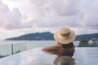 Güneydoğu tropik konseptinde yaz tatili. Arkadan bakan Asyalı kadın, açık havuz tatil köyü deniz manzarasında rahatlıyor. Hasır şapkalı ve dramatik günbatımı gökyüzüyle. Phuket Adası, Tayland