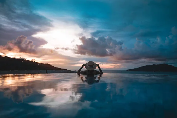Güneydoğu tropik konseptinde yaz tatili. Arkadan bakan Asyalı kadın, açık havuz tatil köyü deniz manzarasında rahatlıyor. Hasır şapkalı ve dramatik günbatımı gökyüzüyle. Phuket Adası, Tayland