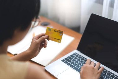 İnternetten alışveriş ve ödeme konsepti. Genç yetişkin bir kadın, maaş bordrosu için kredi kartını elinde tutarak evde kalıyor. Ev ofisi bilgisayarlı çalışma masası.