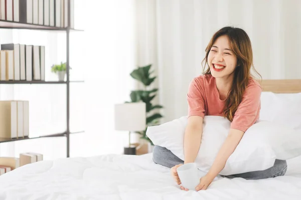 幸せな顔と歯で笑顔でベッドに座って肖像若い大人のアジアの女性 ベッドルームでリラックス後目を覚ますで手を保持コーヒーカップ — ストック写真