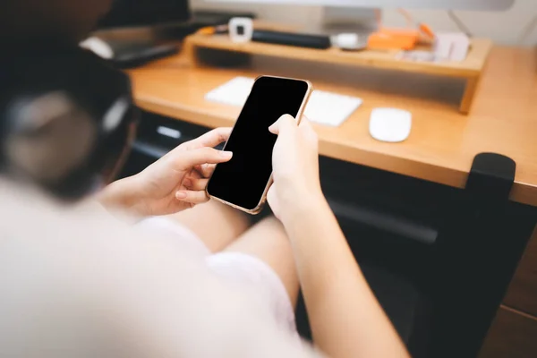 Mock up black blank screen mobile phone. Finger touching on display. Holding by woman hand at home. Workplace office blur background.