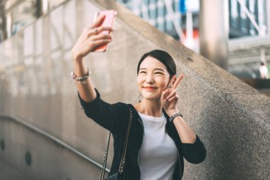 Genç Asyalı bekar kadın cep telefonu selfie 'si ve video görüşmesi kullanıyor. Gülen surat. Şehir hayatı çalışan insanlar. Gündüz dışarıda arka plan gürültüsü.
