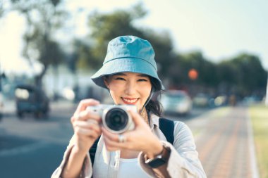 Attractive happy smile young adult asian woman traveller wear blue hat and backpack. Using camera for photo journey. People traveling in city lifestyle on day. Staycation summer trip concept.