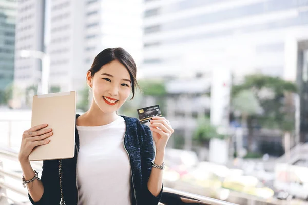 Business people shopping via online application media concept. Happy smile young adult asian woman consumer using creadit card and digital tablet. City on day background with copy space.