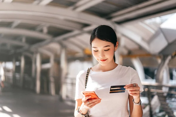 Business people shopping via online application media concept. Happy smile young adult asian woman consumer using creadit card and smartphone. City on day background with copy space.