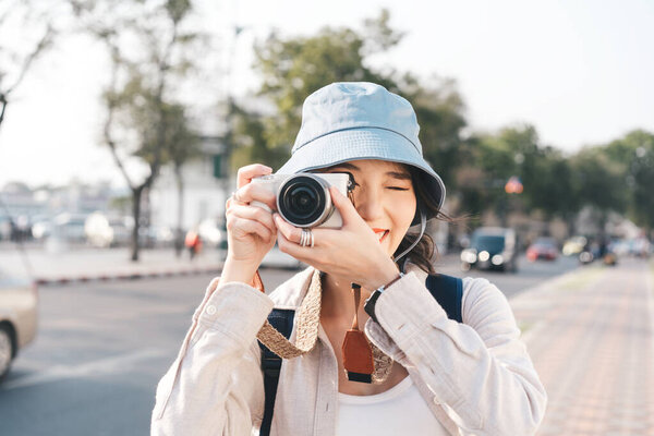 Attractive happy smile young adult asian woman traveller wear blue hat and backpack. Using camera for photo journey. People traveling in city lifestyle on day. Staycation summer trip concept.