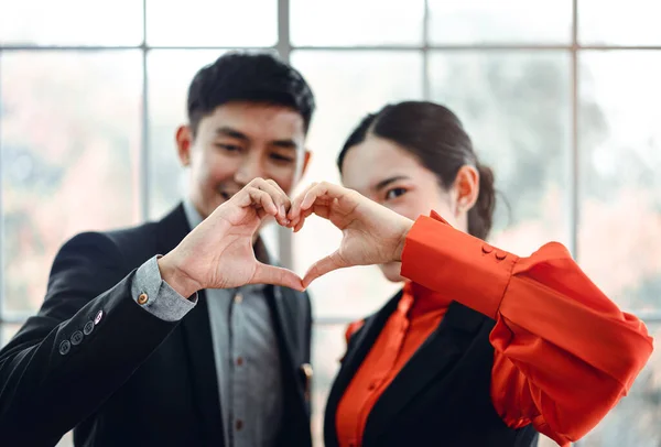 Business smile adult asian man and woman love couple in office concept. They hand use heart gesture. Background in workplace window light with copy space.