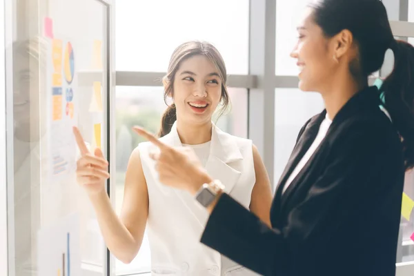 Southeast asian Business people group coworker working in office concept. Happy smile two young adult woman wearing suit talking about strategy on glass board with window day light.