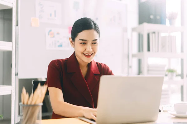 东南亚商人在办公室工作的概念 年轻的女企业家穿着红色西服 用手提电脑在有窗户灯的桌子上 — 图库照片