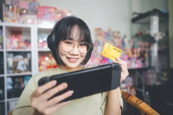 オタクスタイルの若い大人のアジアのゲーマーの女性は眼鏡を着用し ヘッドフォンはハンドヘルドオンラインゲームをプレイします 競争に勝つために支払うためのクレジットカードを使用して — ストック写真