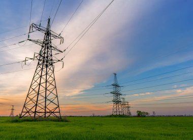 Yüksek voltaj hatları ve elektrik direkleri yeşil tarım alanında canlı ve doygun günbatımı gökyüzüne karşı.