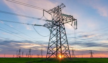 Yüksek voltaj hatları ve elektrik direkleri doymuş günbatımı gökyüzüne karşı yeşil tarımsal bir alanda.