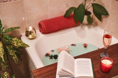 Çiçek yapraklarıyla banyo. Kitap, mumlar ve bir bardak şarap. Konforlu banyoda Organik Spa Rahatlaması
