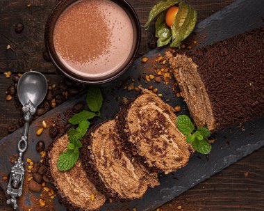 Kütük şeklinde çikolatalı bisküvi, porsiyonlar halinde kesilmiş, çikolata parçacıkları serpiştirilmiş, siyah bir tahtada naneli yapraklarla süslenmiş, kahverengi arka planda bir fincan kakao.