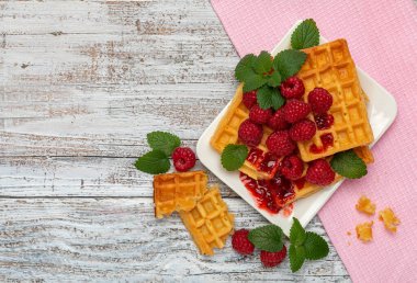 Beyaz bir tabakta Viyana waffle 'ı kırmızı ahududu ve açık ahşap bir masada pembe bir peçetenin üzerinde nane yaprakları. Solda metin için bir yer var.