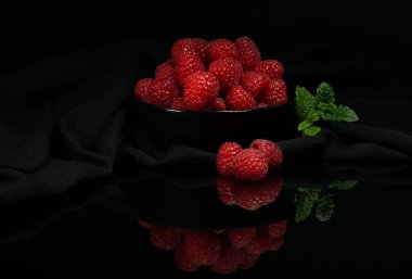 Siyah bir kasede, siyah bir peçetenin üzerinde, siyah bir camla yansıtılmış nane yapraklı olgun kırmızı ahududular.