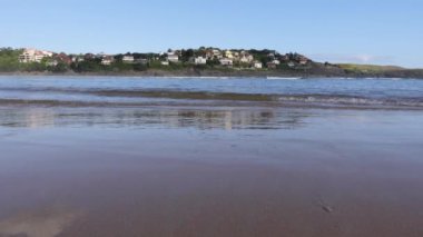 Close-up of the shore of the beach and the waves coming in with the village in the background on a sunny day.