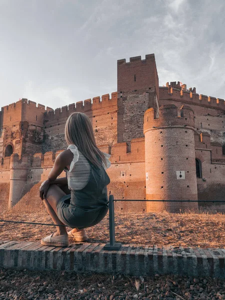 Blonde-haired woman sitting on a wall looking at the castle and its towers with moat in front at sunset.