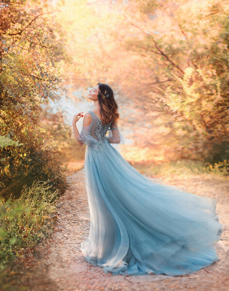 Happy fairy woman princess in light summer blue dress, standing in autumn park orange foliage tree. Lady walks fashion model. fantasy girl goddess. flying long hem of dress on wind. nymph of forest