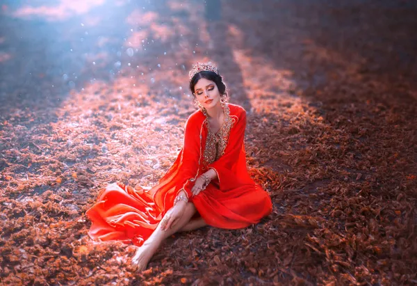 Mujer joven de fantasía en vestido de seda oriental rojo se sienta sobre hojas naranjas caídas. Lady Princesa en corona de oro, rubíes. Reina en estilo del Imperio Otomano. Chica belleza árabe en el fondo otoño naturaleza soleada. —  Fotos de Stock