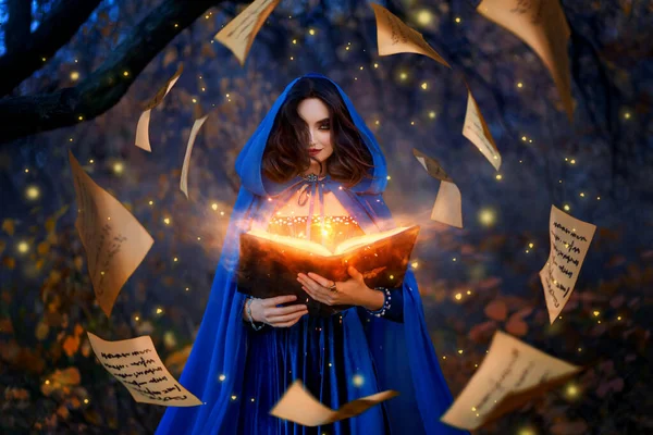 Mulher de fantasia bruxa mágico no capuz detém em mãos livro de mágica, feitiços de luz laranja brilhante, vento dispersa folhas de queda levitação página de papel. Feiticeira. Manto medieval vestido azul traje mágico. — Fotografia de Stock