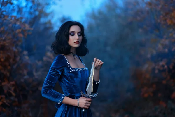 Fantasía mujer medieval guerrera reina sosteniendo daga, cuchillo en las manos. Vestido azul vintage de terciopelo real con perlas, vampiro princesa chica, pelo corto morena. Naturaleza bosque noche de anochecer, temporada de otoño — Foto de Stock