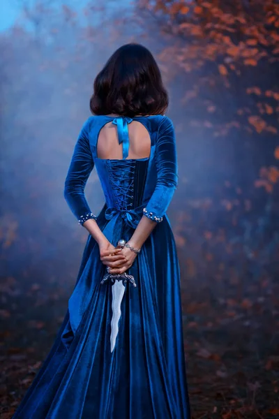 Fantasy warlike medieval woman queen holds metal vintage dagger blade weapon in hands hides behind back. Background forest fog night. Blue velvet luxurious royal dress. Girl princess warrior rear view — Stock Photo, Image