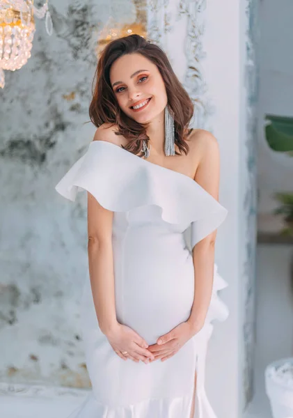 Young beautiful pregnant woman with smile on face, gently hugs embrace her stomach belly with hands. White evening elegant pregnancy dress. Light room classic interior background. Tight wedding dress. — Foto Stock