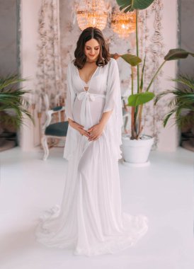 Young beautiful pregnant woman stand in classic room hugs bare belly with hands. White long silk negligee pregnancy dress peignoir, dressing gown. Light room classic background. Gently smiling face