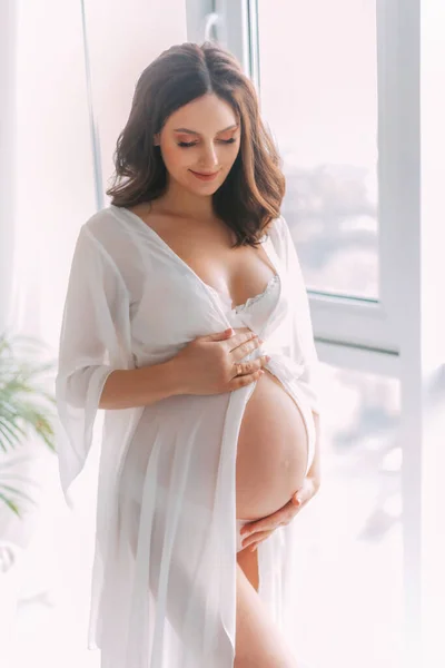 Young beautiful pregnant woman stand near window hugs bare belly with hands. White long silk negligee pregnancy dress peignoir, dressing gown. Light room classic background. Gently smiling face —  Fotos de Stock