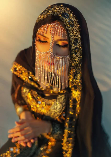 Retrato mulher oriental sentar no deserto de areia ao pôr do sol. Menina rosto escondido pela cabeça do véu de ouro lenço coberto. Noite brilhante estilo árabe make-up, olhos fumegantes. dourado brilhante preto abaya vestido tradicional — Fotografia de Stock