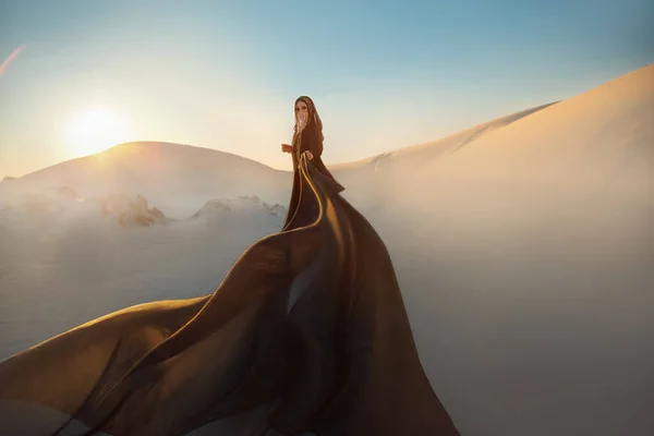 Mystery arabic woman in black long dress stands in desert long train silk fabric fly flytter in wind motion. clothes gold accessories hide face. Oriental fashion model. Sand dunes background sunset. — Foto Stock