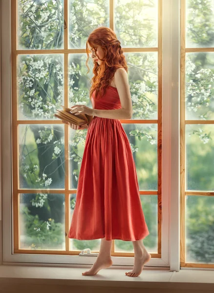 Medieval woman princess holds in hands reads book. Red-haired dreamlike girl stands on window barefoot, view summer green garden trees. Red vintage dress, ball gown. Long red curly hair, pale skin — Foto Stock