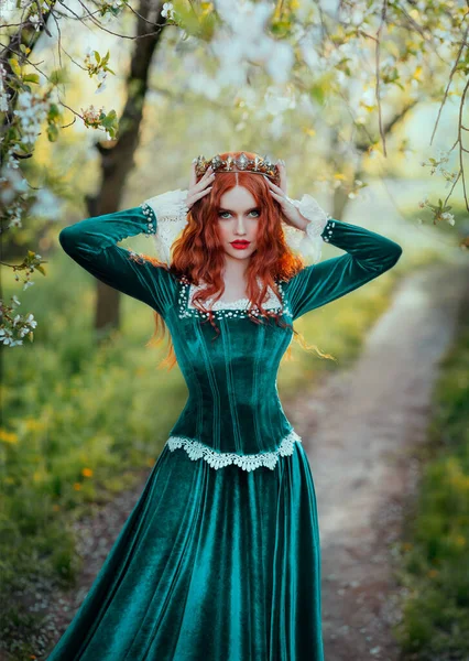 Portrait fantasy red-haired woman medieval queen touches with hands straightens golden crown on head. Girl redhead princess. Green vintage long dress curly hairstyle. Summer nature garden forest trees — Fotografia de Stock