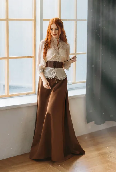 Red-haired woman in vintage dress stands at large classic window waiting love. Clothing costume countess old style white blouse, brown long skirt. Curly red hair. Redhead girl princess 1800s stylish — Foto Stock