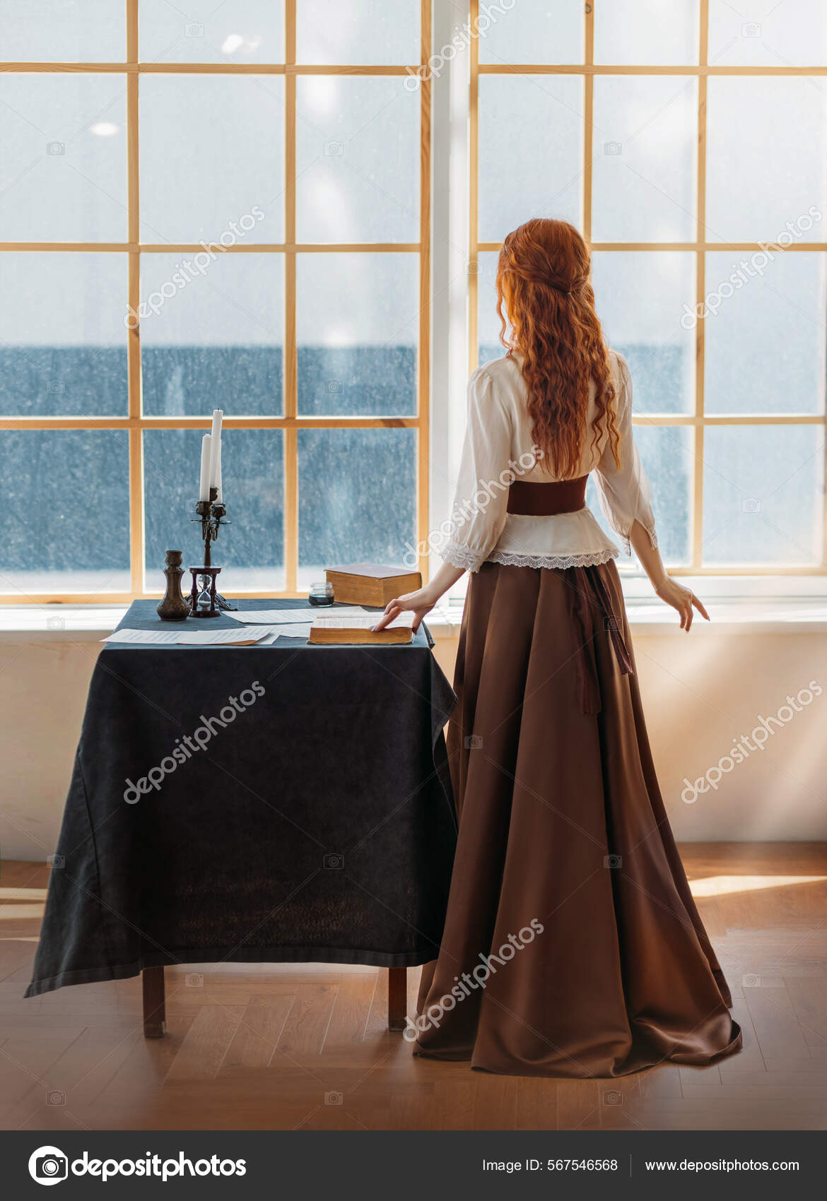 Red-haired woman in vintage dress stands looks at classic window waiting  love. Clothing costume countess old style white blouse, brown long skirt.  Curly red hair. Redhead girl princess back rear view Stock