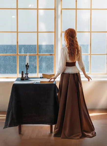 Red-haired woman in vintage dress stands looks at classic window waiting love. Clothing costume countess old style white blouse, brown long skirt. Curly red hair. Redhead girl princess back rear view — Foto Stock