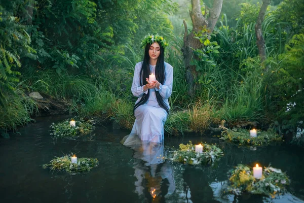 Elinde mum tutan fantezi pagan cadı Slav kadın. Kehanet yaz geleneği. Deniz kızı perisi nehir kıyısında oturuyor. Beyaz, klasik gecelik elbisesi. Yaz doğası yeşil çimen ağaçları. Uzun saçlı.