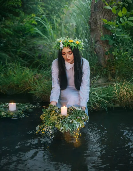 Σλάβα γυναίκα κρατά φυτικό στεφάνι στα χέρια βάζει στο νερό, κεριά επιπλέουν. Φαντασία κορίτσι νύμφη Λευκό μακρύ φόρεμα. Θερινό πράσινο γρασίδι όχθη του ποταμού. Έννοια ειδωλολατρική αργία του Ιβάν Κουπάλα μαντεία παρθένες — Φωτογραφία Αρχείου