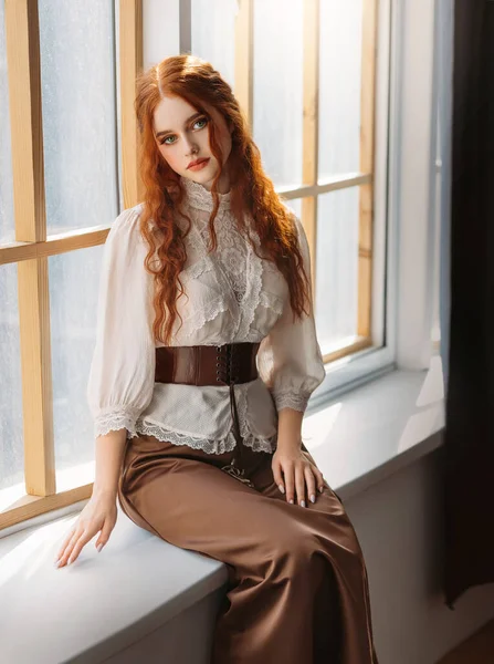 Red-haired woman in vintage dress sits on large classic window waiting love. Clothing costume countess old art style white blouse, brown long skirt. Curly red hair. Redhead girl princess 1800s stylish — Foto Stock