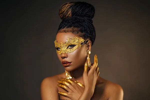 Portrait sexy african woman face close up in golden venetian carnival mask. Girl afro fashion model perfect skin, golden evening makeup, glitter diamond stones lip gloss, hands in gold liquid paint