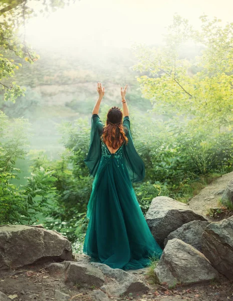 Fantasy red hair woman queen prays in summer forest hands raised to sky. Girl in long elegant royal vintage elven green dress. wide long sleeves silk fabric flying in wind. Art photo bare open back