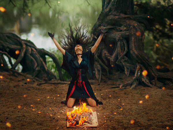Fantasy female pagan witch creating magic casts spell dancing ritual dance near burning hearth of fire, waves head arms hands raised to sky, long black hair flying in wind motion. Night forest trees.