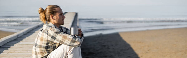 Side view of longhaired man holding cup on wooden pier on beach in Italy, banner 