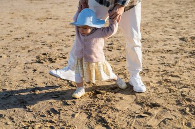 Baby girl in panama hat holding hands of father while walking on beach  clipart