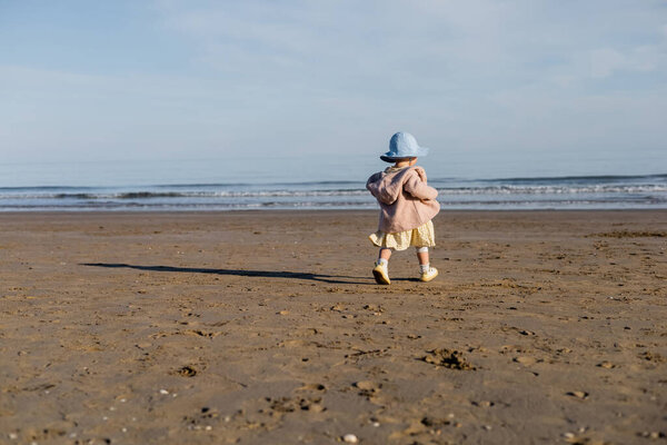 Back view of baby girl in panama hat walking on sandy beach in Italy 