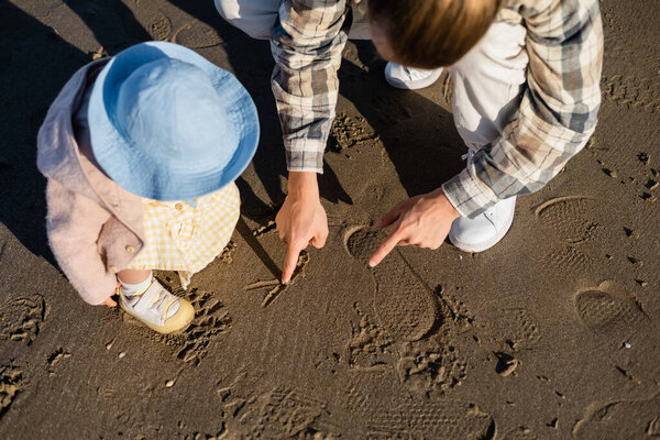 Overhead view of dad drawing on sand near baby in panama hat on beach 